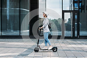 Side view of a young businesswoman riding electric push scooter. Female in cycling helmet driving electrical scooter against