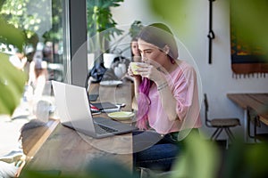 Side view of young businesswoman sitting at table in coffee shop. On table cup of coffee and laptop. In background white