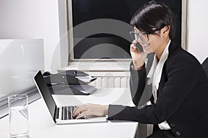 Side view of young businesswoman using laptop and cell phone at desk in office