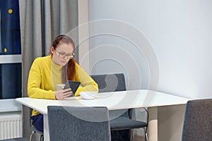 Side view of the young caucasian woman in yellow knitted sweater seats at the table in modern light interior and reading an