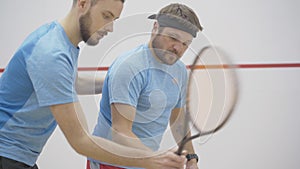 Side view of young coach correcting posture and gripping technique of bearded tattooed sportsman playing squash in gym