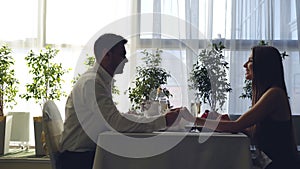 Side view of young couple sitting together at table in restaurant and talking holding hands. Romantic relationships