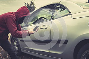 Side view of young criminal in black balaclava and hoodie holding smartphone and trying to interact with car by mobile