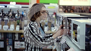 Side view of a young curly haired woman in plaid shirt chooses a microwave oven in a consumer electronics supermarket