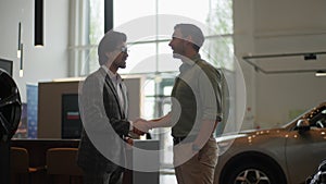 Side view of young customer male thanking stylish dealer male wearing suit and eyeglasses for helping to choose car to