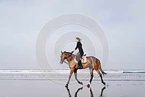 side view of young female equestrian riding horse on sandy