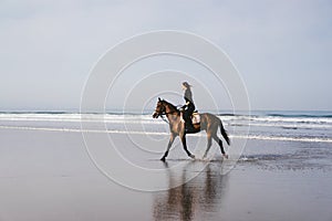 side view of young female equestrian riding horse on sandy