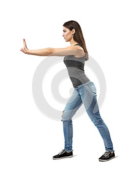 Side view of young fit woman in gray top and blue jeans, with long dark hazelnut hair, posing as if pushing something