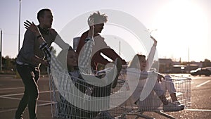 Side view of a young friends having fun outdoors on shopping trolleys. Multiethnic young people racing on shopping carts