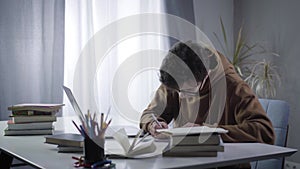 Side view of young genius in eyeglasses typing on laptop keyboard and writing in workbook. Portrait of absorbed