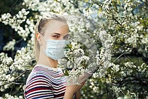 Side view of young girl in medical mask looking at camera on background of blossoming apple tree on summer day.