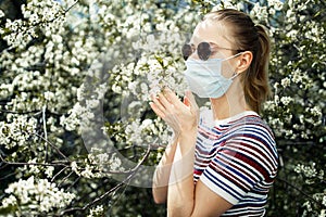 Side view of young girl in medical mask and sunglasses on background of blossoming apple tree on summer day.