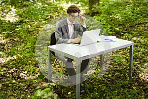 Side view of Young handsome business man working at laptop at office table in green park. Business concept.