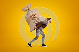 Side view of young handsome man in casual clothes carrying huge heavy sack with dollar symbol on.