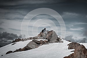 Side view of a young hiker man resting on a rock at the top of a snowy mountain during sunrise