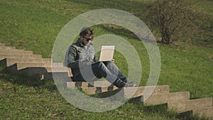 Side View of Young Hipster Man In Sunglasses Sitting On The Stairs Using Laptop