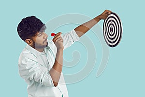 Side view of young Indian man holding target and aiming little arrow at bullseye