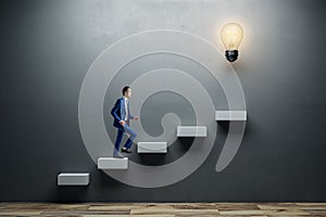 Side view of young man climbing illuminated light bulb stairs to success on concrete wall background in interior. Financial growth