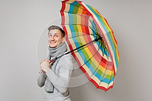 Side view of young man in gray sweater, scarf hold colorful umbrella looking up on grey background. Healthy