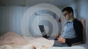 Side view a young man is lying on a bed and working on a laptop, typing on a keyboard, working at night, remote work