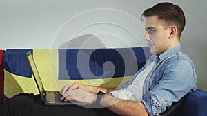 Side view a young man is lying on the sofa and working on a laptop, typing on the keyboard, remote work from home.