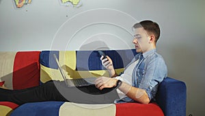 Side view a young man is lying on the sofa and working on a laptop, typing on the keyboard, remote work from home.