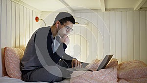 Side view a young man is sitting on the bed and working on a laptop, plugging in headphones and typing on the keyboard