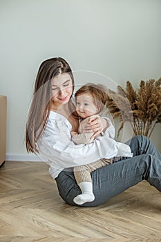 Side view of young mother sitting on floor at home and holding little beautiful baby daughter
