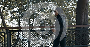 Side view of young pretty muslim woman running in park. Energetic female person in hijab and sports suit jogging