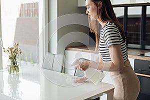 Side view of young pretty woman standing in kitchen, spraying on white table with vase from pulverizer, wiping with rag.