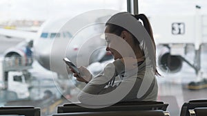 Side view young pretty woman using smartphone in waiting room in airport background of plane in window