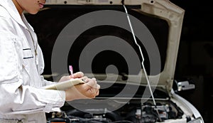 Side view of young professional mechanic in uniform writing on clipboard against car in open hood at the repair garage. Insurance