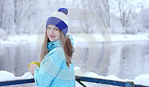 Side view of young smiling woman with a cup of hot tea or coffee on snowy winter waterside.