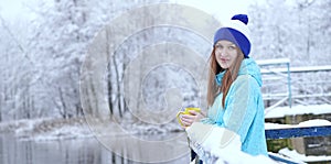 Side view of young smiling woman with a cup of hot tea or coffee on snowy winter waterside.