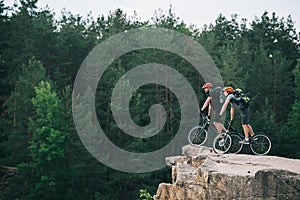 side view of young trial bikers standing on rocky cliff with blurred pine forest on background and looking