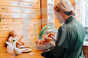 Side view of young woman craftsman decorating newborn baby doll with sponge while sitting in workshop at home.