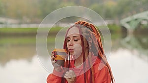 Side view of young woman drinking coffee. Female with redheads dreadlocks with a cup of coffee or tea on background of