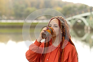 Side view of young woman drinking coffee. Female with redheads dreadlocks with a cup of coffee or tea on background of