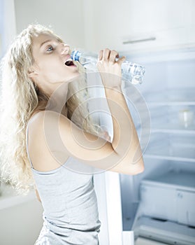 Side view of young woman drinking water from bottle by refrigerator