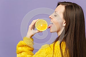 Side view of young woman in fur sweater hold licking half of fresh ripe orange fruit isolated on violet pastel