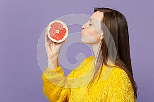 Side view of young woman in fur sweater hold sending air kiss to half of fresh ripe grapefruit isolated on violet pastel