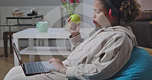 Side view of young woman in headphones biting chewing healthful apple messaging online on laptop. Confident African