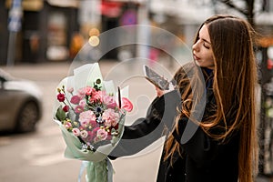 Side view of young woman holding delicate flower arragement wrapped in mint color paper and takes shot on iphone