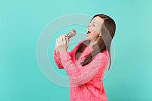 Side view of young woman in knitted pink sweater with closed eyes hold in hand, sing song in microphone isolated on blue