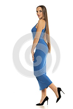 Side view of a young woman in long transparent blue dress walking on a white background