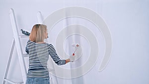 Side view of young woman paints with brush roller plastered wall in white color with plastic tub