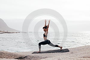 Side view of young woman practicing yoga at sunset by bay. Full length of female in Warrior pose on mat outdoors