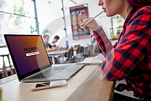 Side view of a young woman sitting in cafe, hand typing laptop computer with password login on screen.