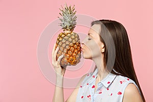 Side view of young woman in summer clothes hold kissing fresh ripe pineapple fruit isolated on pink pastel wall