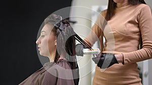 Side view young woman talking with hairdresser straightening hair with flat iron in beauty salon. Caucasian female